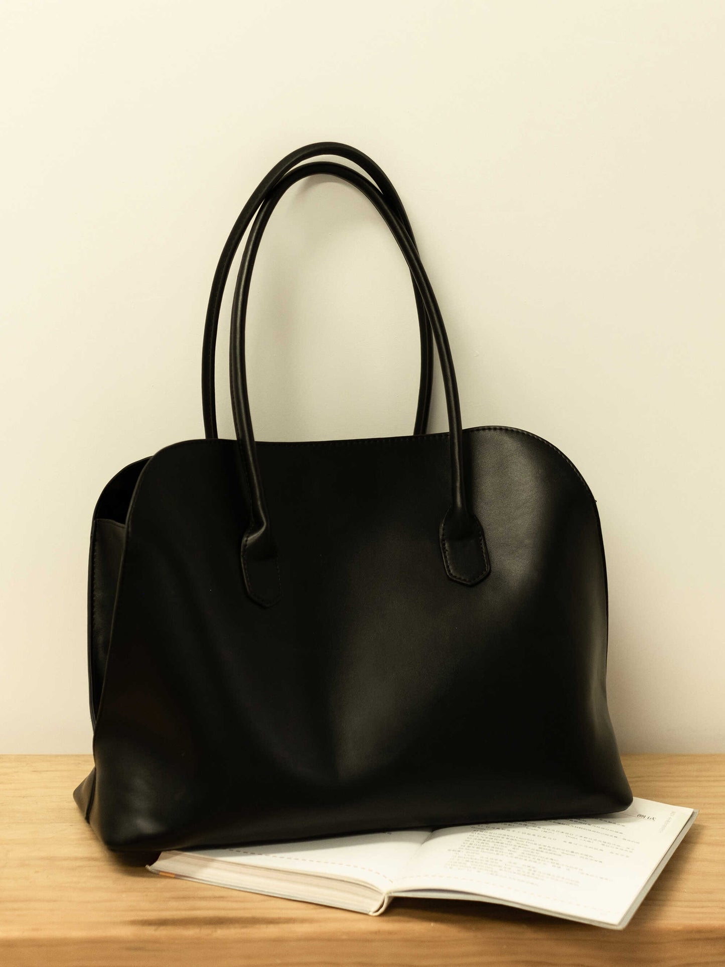 A black leather shoulder bag resting on a book atop a wooden table.