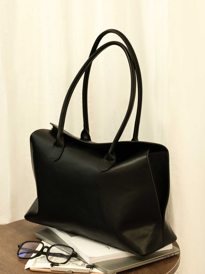 A black leather Alma bag rests on a pile of books and a laptop atop a wooden table, next to a pair of glasses.