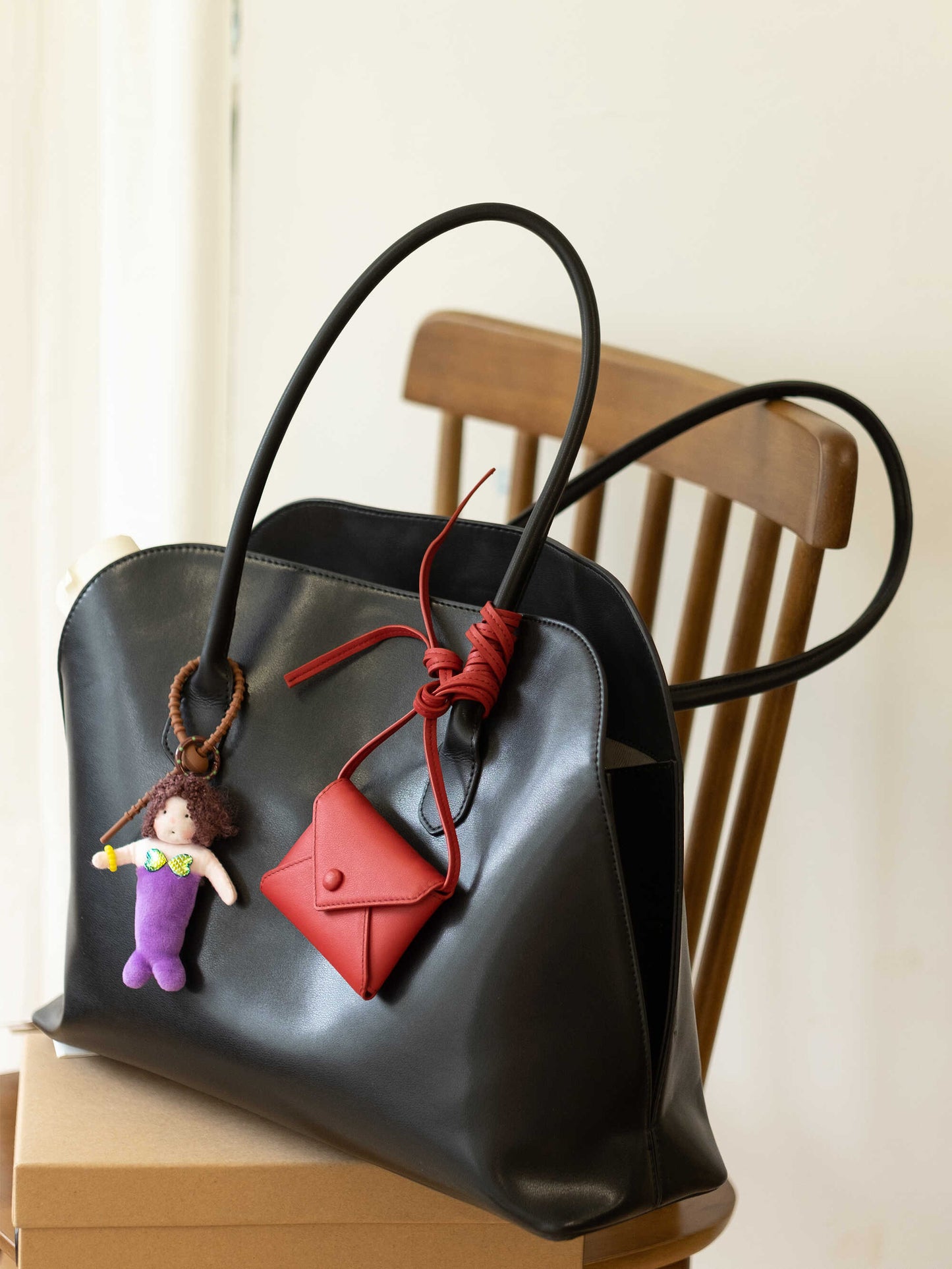 A black leather Alma bag with a mermaid charm and a red envelope purse tied to the handle rests on a paper box atop a wooden chair.