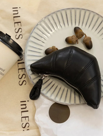 A top view of the croissant-shaped black purse lies flat on a white plate, next to a coffee cup and four pinecones.