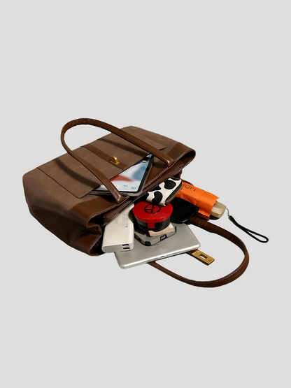 A brown work tote bag lies on a white background, stuffed with items such as an iPad, makeup, iPhone, and an umbrella, showcasing its spacious design.