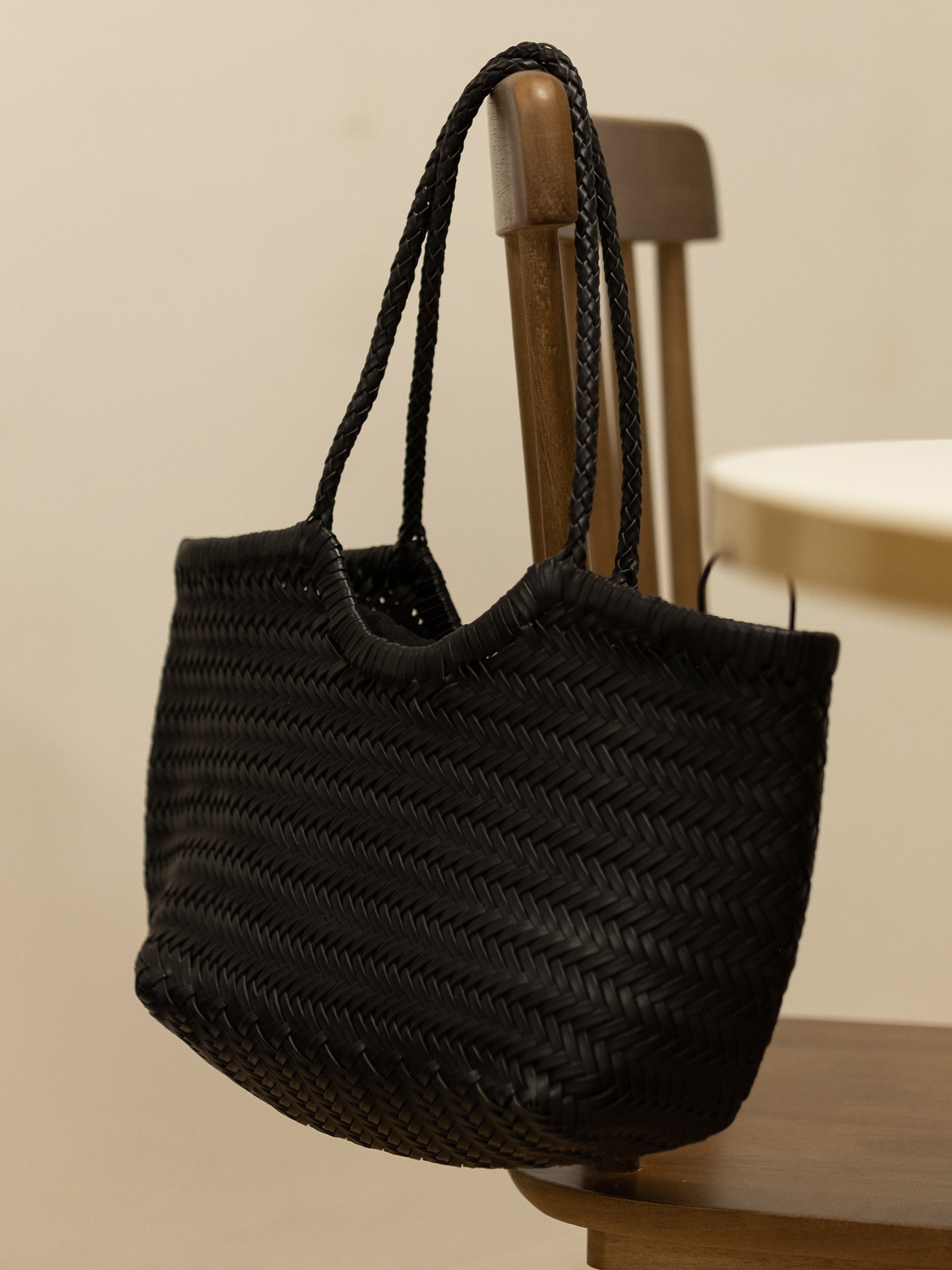 A black woven leather tote bag hanging on the back of a wooden chair, showcasing its functionality and fine woven technique.