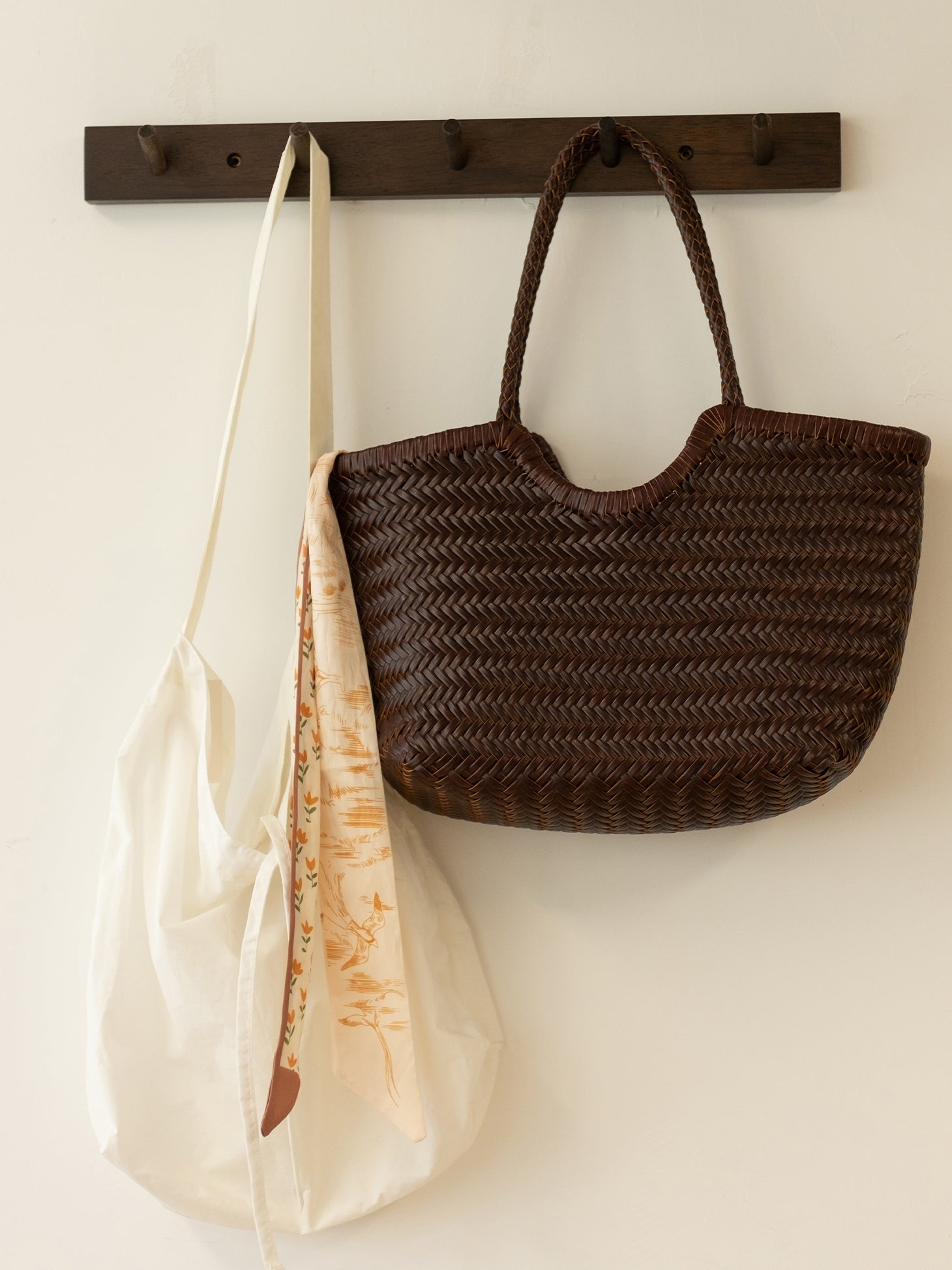 A brown woven leather tote bag hanging against a white wall, next to a white canvas bag, highlighting its spacious and functional design.