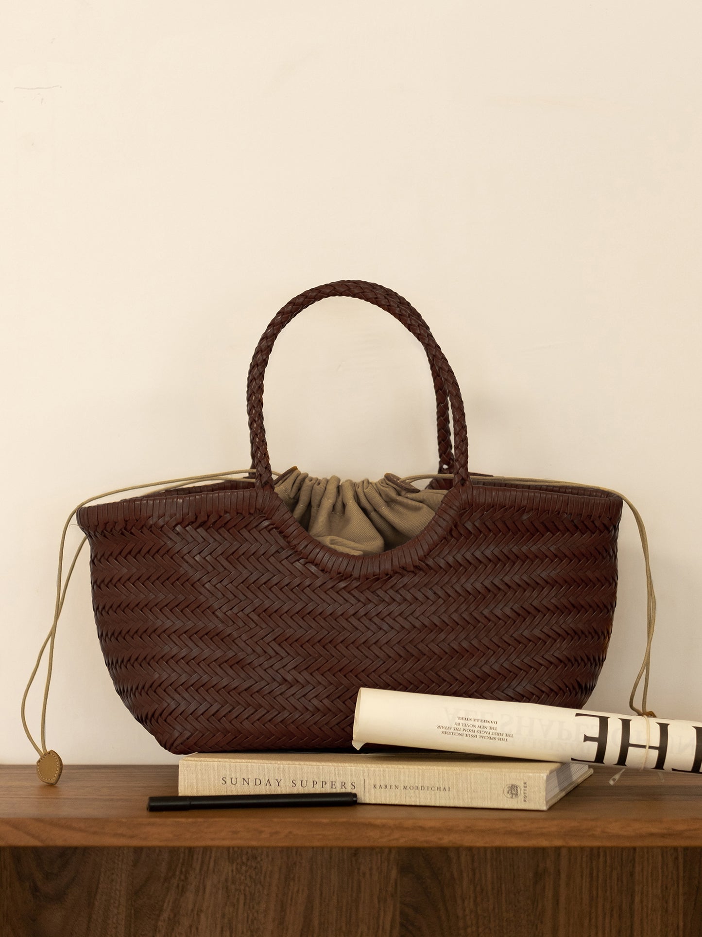 A front view of a brown woven leather tote bag hanging with a matching color nested bag, resting on a book atop a wooden table.