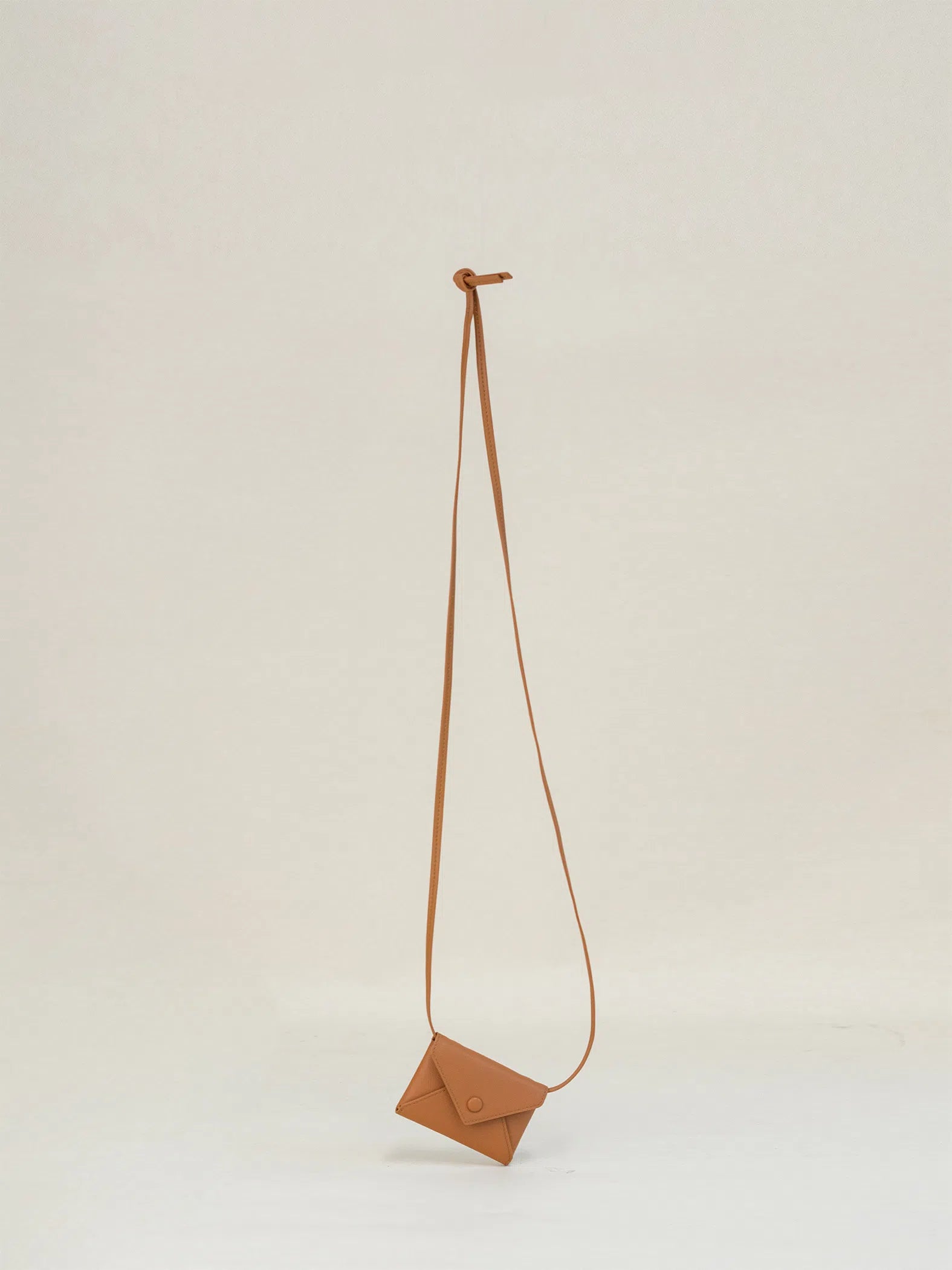 An envelope-shaped creamy apricot crossbody wallet, made from real leather and featuring a long strap, hangs against a white background.