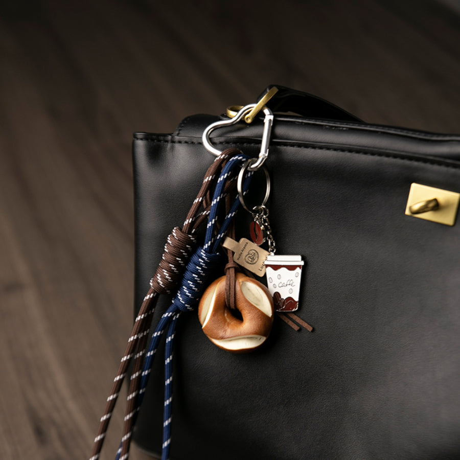 A closer look of a set of unique bag charms, including a coffee cup, a pretzel, and blue and brown braided cords, hanging from a black leather bag against a wooden background.