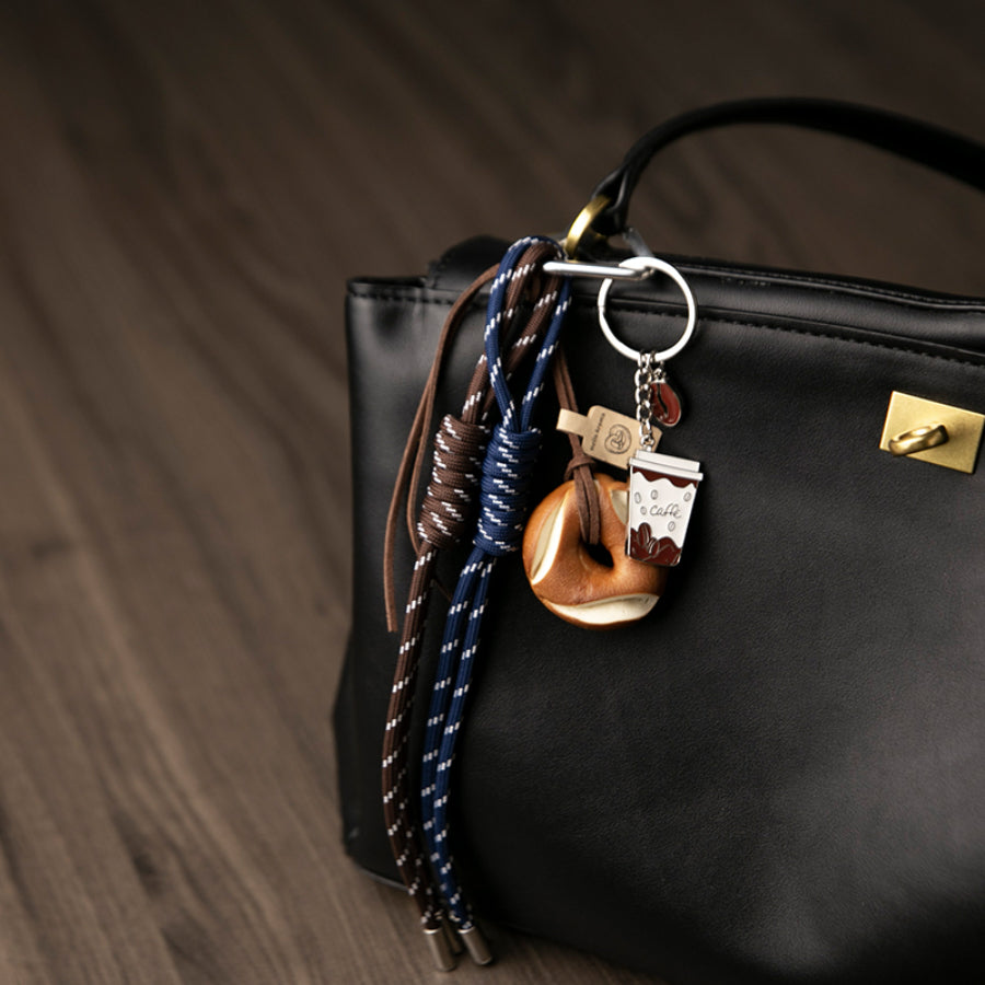 A set of unique bag charms, including a coffee cup, a pretzel, and blue and brown braided cords, hanging from a black leather bag against a wooden background.