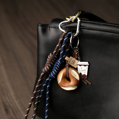 A even closer look of a set of unique bag charms, including a coffee cup, a pretzel, and blue and brown braided cords, hanging from a black leather bag against a wooden background.