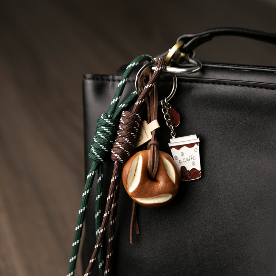 A closer look of a set of unique bag charms, including a coffee cup, a bagel, and green and brown braided cords, hanging from a black leather bag.