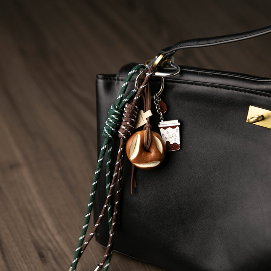 A set of unique bag charms, including a coffee cup, a bagel, and green and brown braided cords, hanging from a black leather bag, against a wooden background.