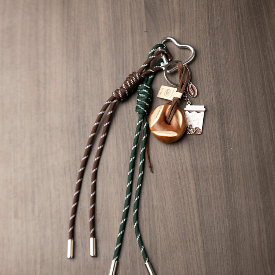 A set of unique bag charms, including a coffee cup, a bagel, and green and brown braided cords, rests on a wooden background.