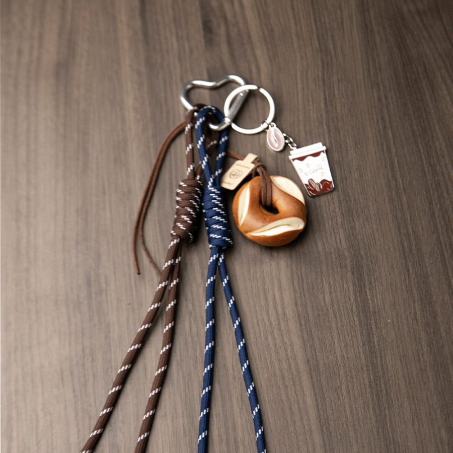 A set of unique bag charms, including a coffee cup, a bagel, and blue and brown braided cords, rests on a wooden background.